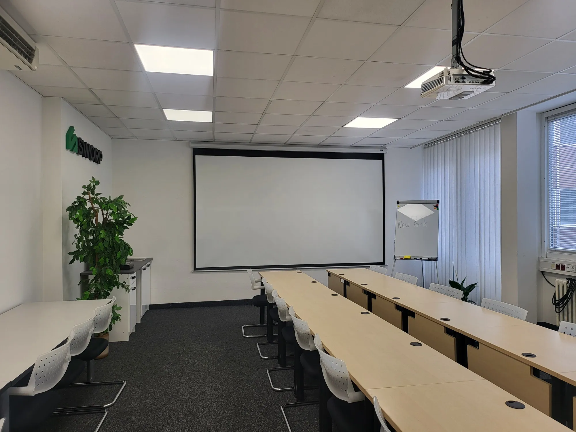 Modern conference room with long tables, white chairs, a projector, a blank screen, a flip chart with 'New York' written on it, and a potted plant near the wall.