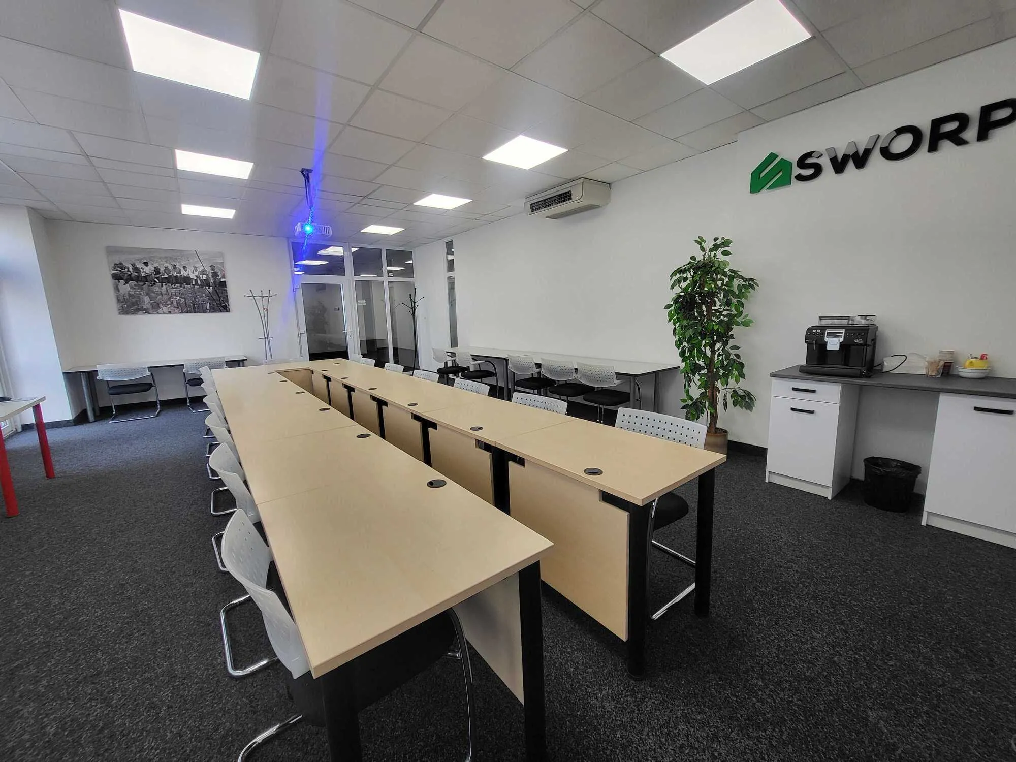 Modern office conference room with long wooden tables, white chairs, overhead lights, and a coffee machine on a countertop.