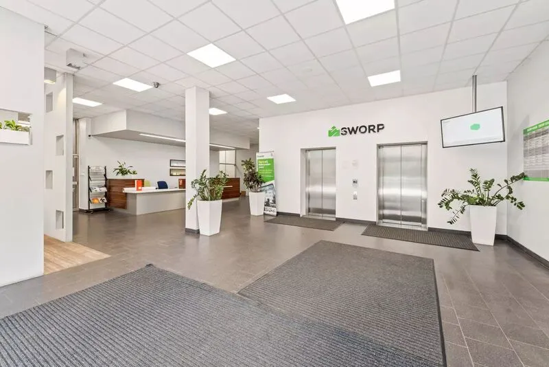 Modern office lobby with two stainless steel elevators, a reception desk, and several potted plants.
