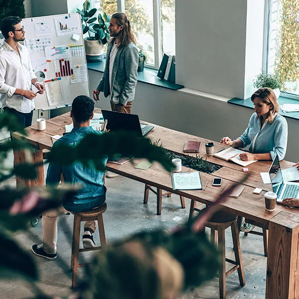Four colleagues collaborating in a modern office with laptops, charts on a whiteboard, notebooks, and coffee cups on a wooden table near large windows.