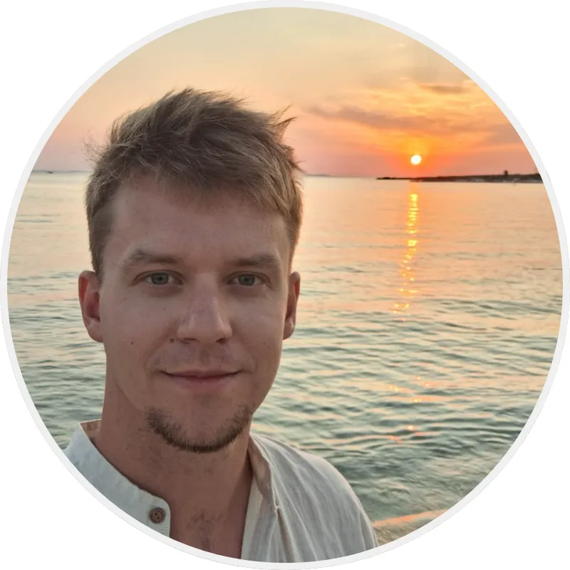 Muž s krátkými vlasy a světlou košilí se usmívá u moře při západu slunce.