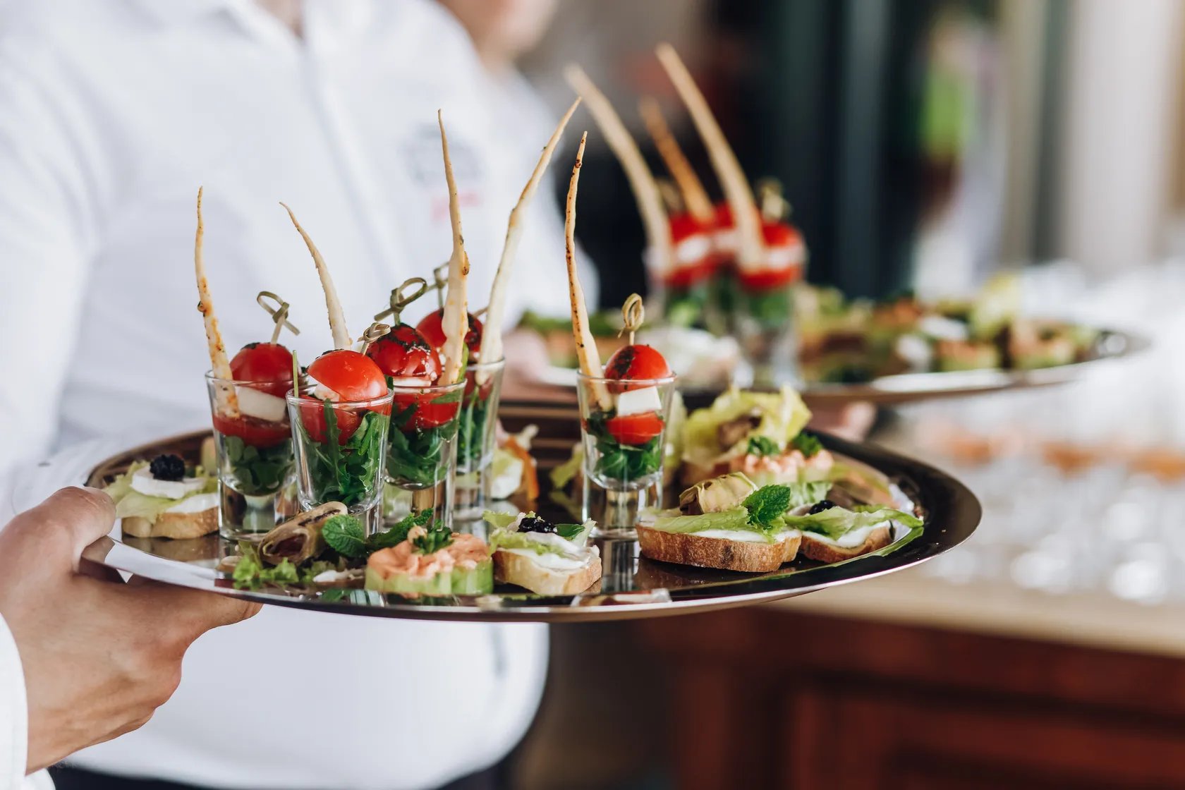 Waiter holding a tray with assorted appetizers including salad in shot glasses and small open-faced sandwiches.